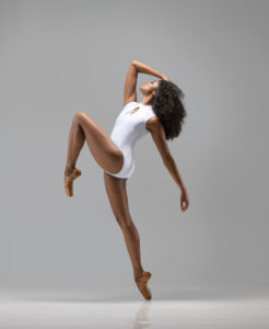 A black woman in a white leotard with her knee up feet pointed and head leaning back with one arm up to her head. She's lit in a professional and clean manner allowing for a feint reflection to extended downwards from her base foot
