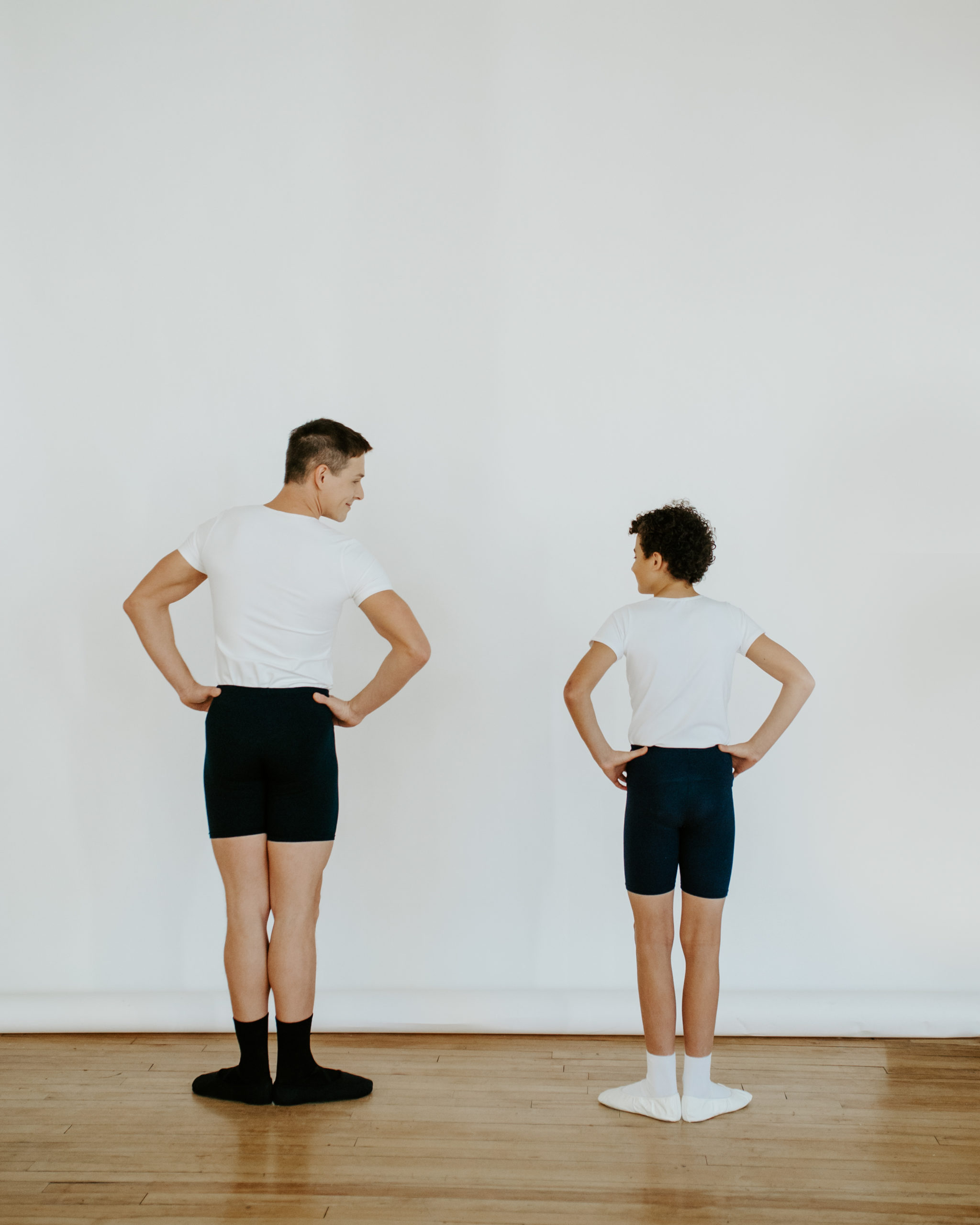 IMG_8007-Edit-ssf A man and a boy standing beside one another both in a white shirt and black shorts with heels together and toes outturned. The man looks over at the boy and the boy over at the man. Both have their hands on they hips