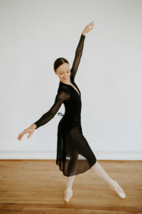 A woman in a black leotard with a flowing cardigan like sheer cover stands in center frame with arms flowing from low to high and one leg across the other with one foot pointing at the viewer and another pointing in the direction of the legs movement