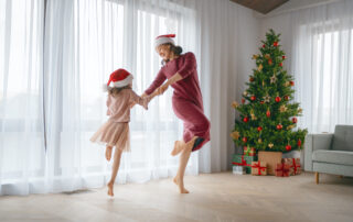 Merry Christmas, Happy Holidays. Cheerful mom and her cute daughter girl are dancing. Parent and little child having fun near tree indoors.\ family celebrating Christmas