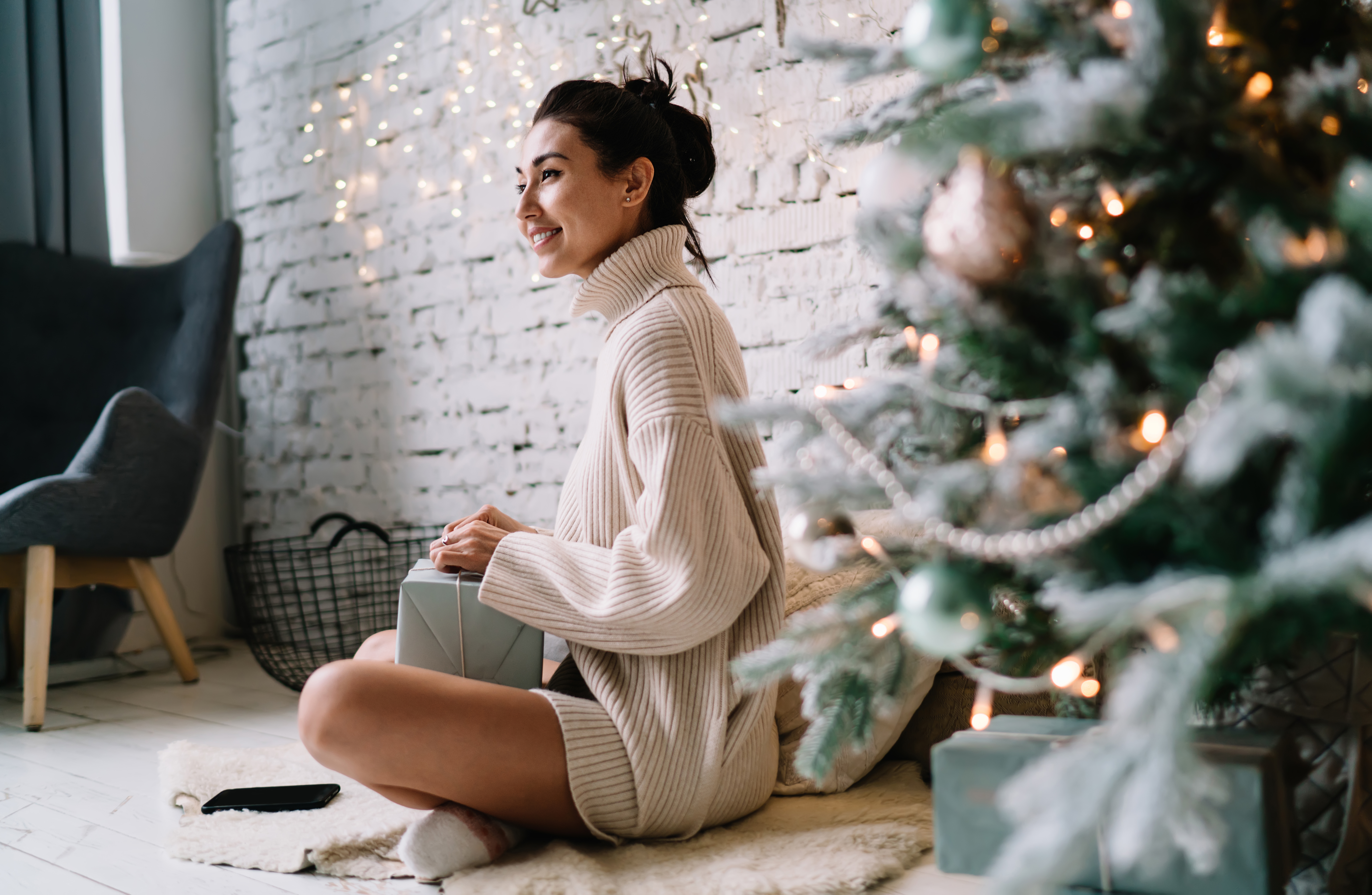Chinese girl sittng on furry carpet near Christmas tree and opening gift present at home. Christmas and New Year holidays. Young beautiful woman looking away. Domestic lifestyle. Modern apartment